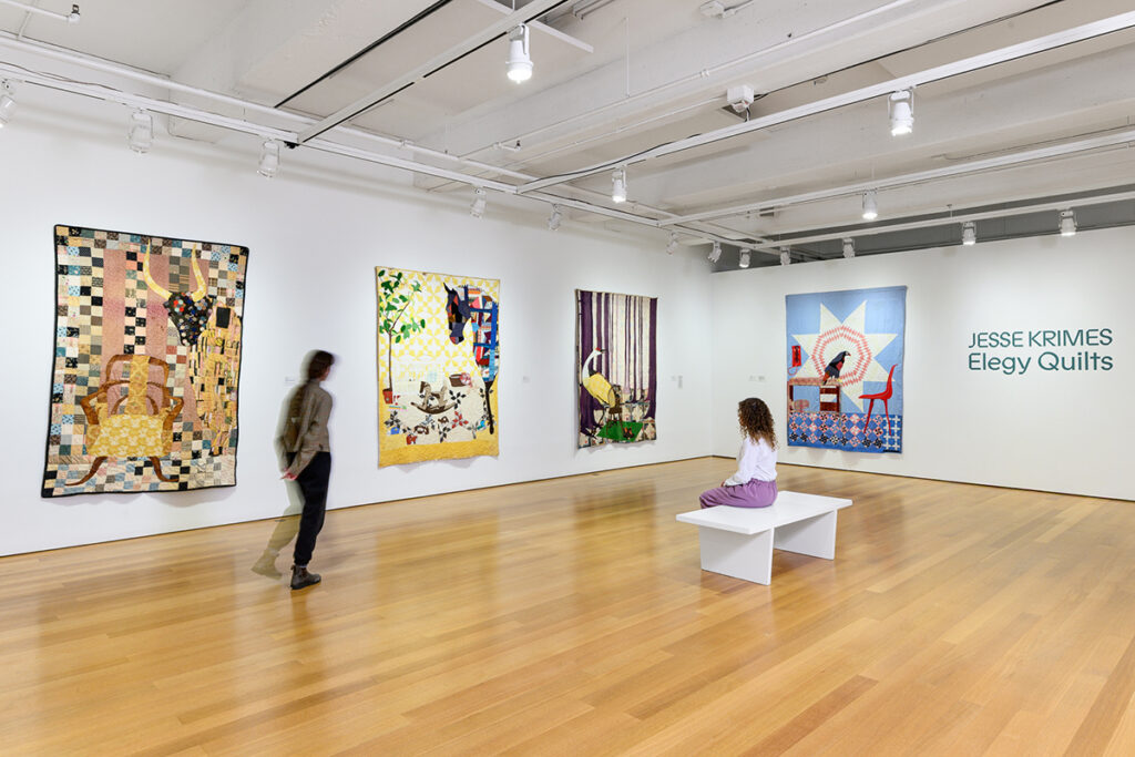Two museum visitors view a series of large quilts in a gallery setting.