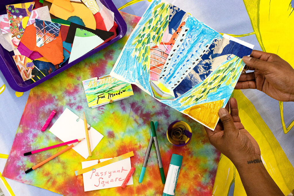 A person holding a folded out collaged drawing featuring different areas of colors, shapes, and patterns. There are other samples on the table below, suggesting this sample is an abstracted map.