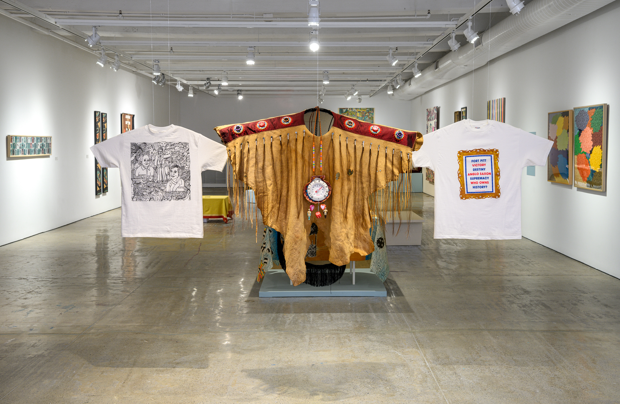 A gallery installation showing three garments, including two t-shirts and a Native American shroud.