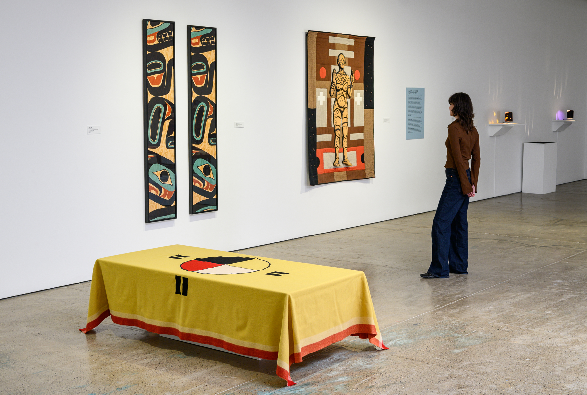 A gallery view of three artworks, including a blanket overlaying a low-rise box, two vertical scarves, and a blanket featuring a ceramic goddess figure.