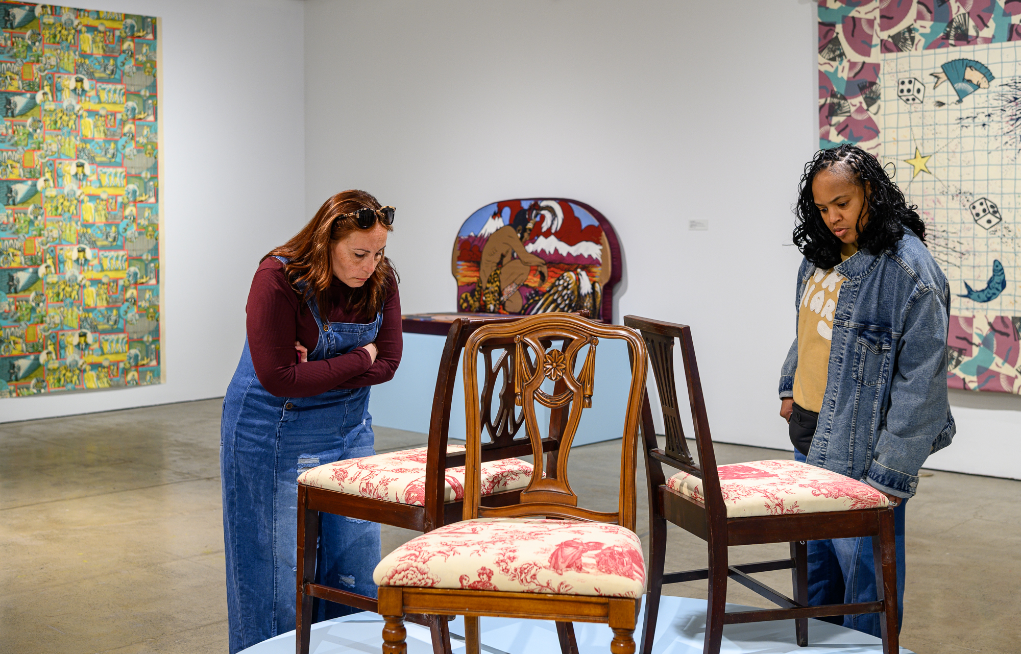 A picture of two women looking at a set of three chairs upholstered with detailed imagery. Behind them are more artworks with patterns and illustrations.