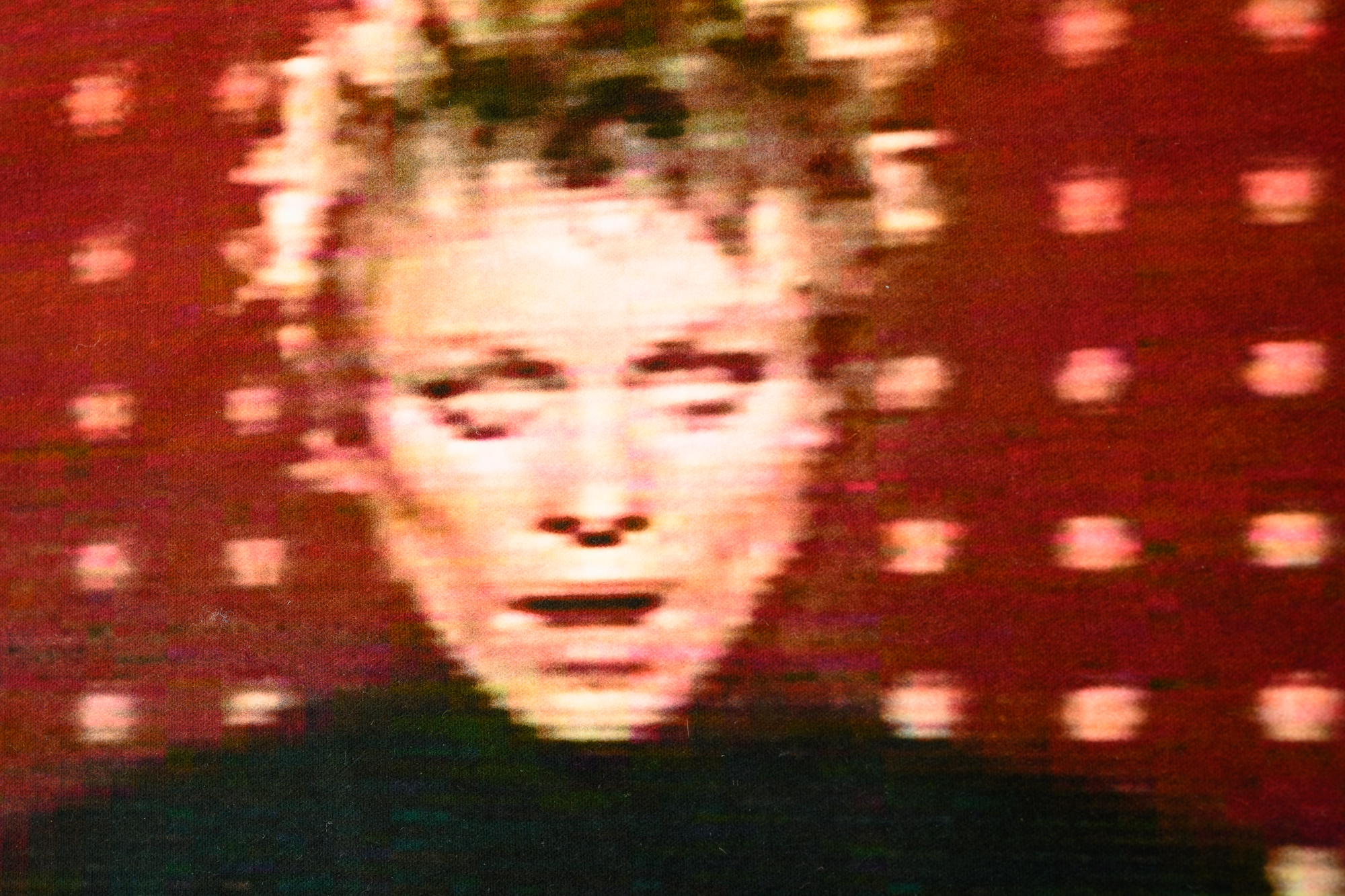 A detail of a rug artwork by Laurie Anderson that is a film still from her music video, O Superman. The focus is on the artist's face, a white woman with short blonde hair, staring at the viewer in front of a red environment. The pile of the rug mimics the film grain, pixelating her image.