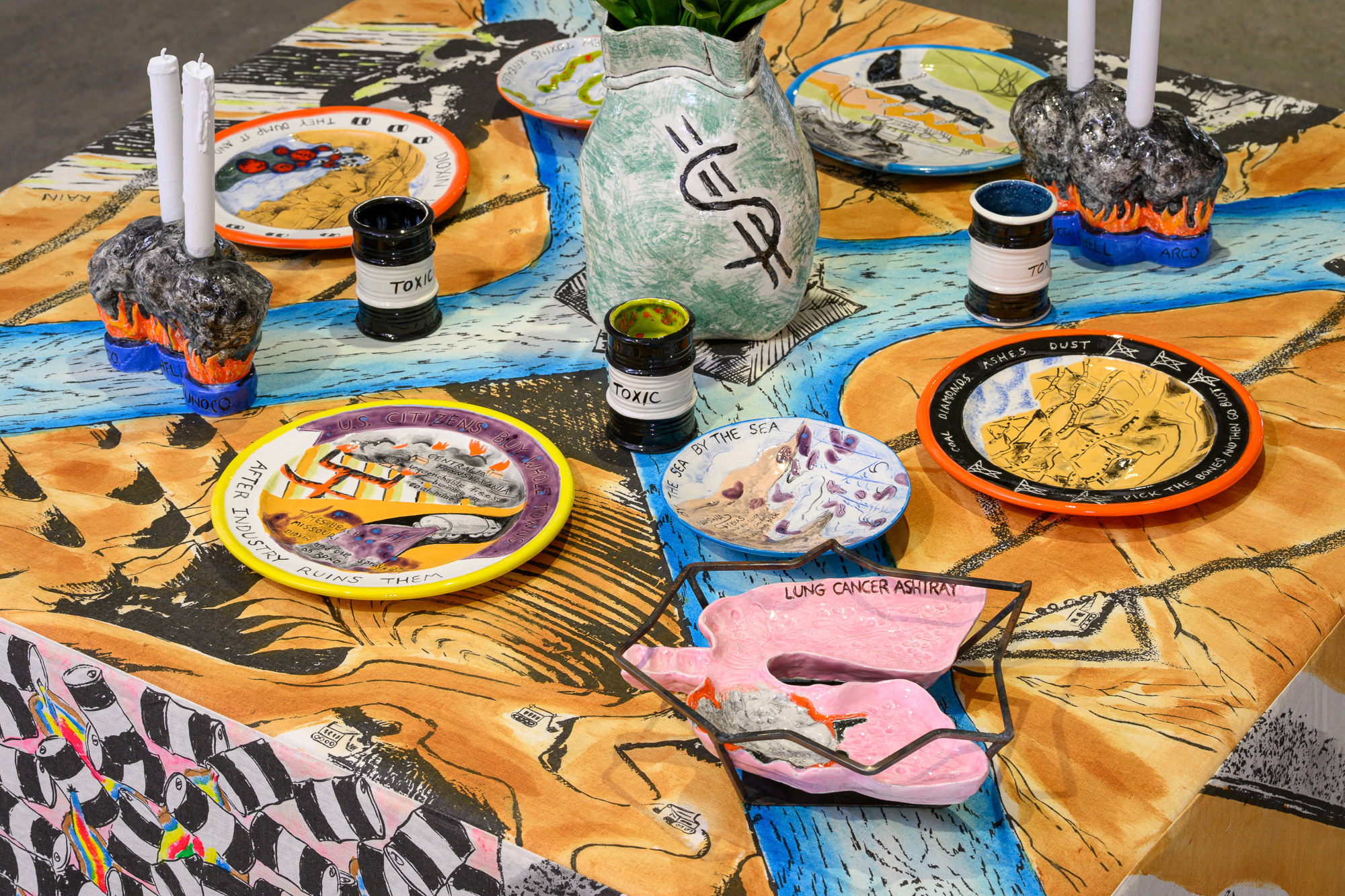 A tablescape showing an arrangement of ceramic plates, candle holders of smoky oil plumes, cups that resemble toxic waste barrels, a vase with a dollar sign, and an ashtray resembling cancerous lungs.