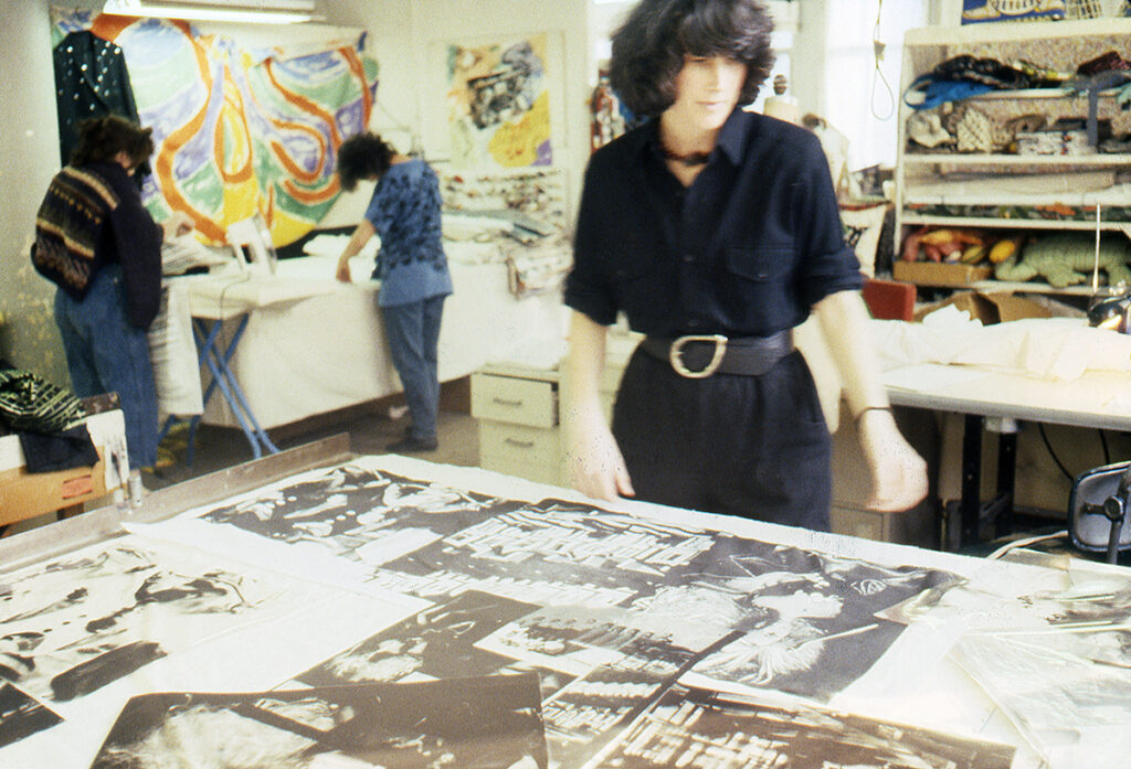A white woman stands over a large table in an art studio. She is working on arranging various black and white pictures in a composition. In the background, two other artists stands over a table ironing.