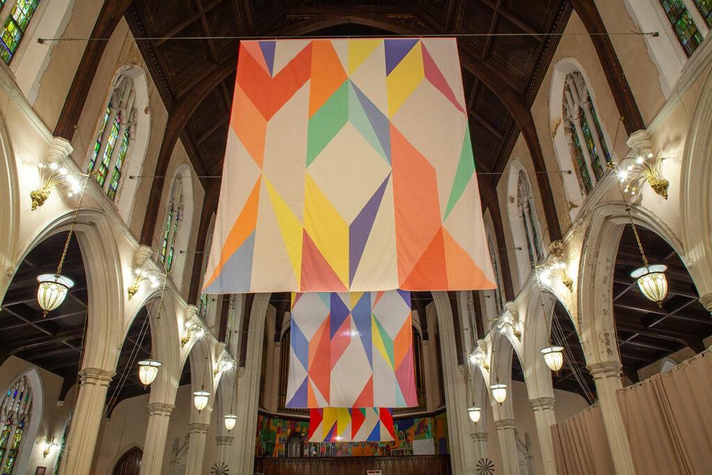 A photo of a church sanctuary with three large banners suspended from the vaulted ceiling. The rectangular designs feature abstract diamond and chevron shapes of various colors.
