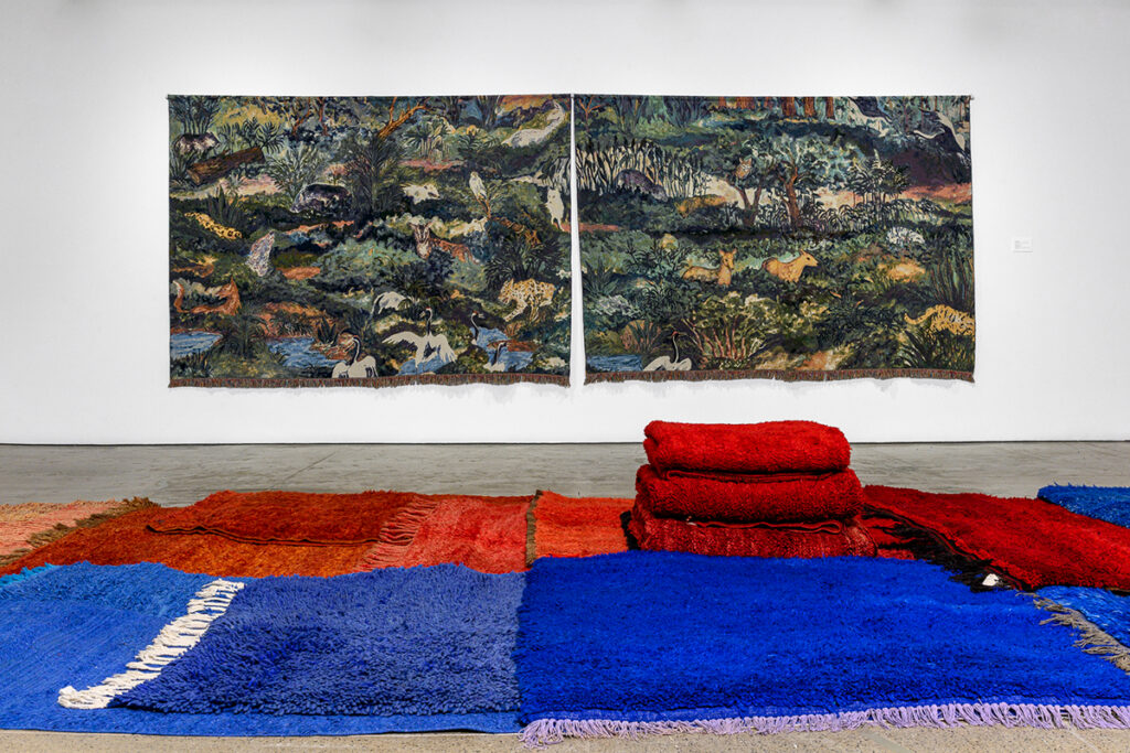 A diptych of two long tapestries featuring a forested scene of woodland creatures hangs on a gallery wall while a stack of rugs stretches across the floor.