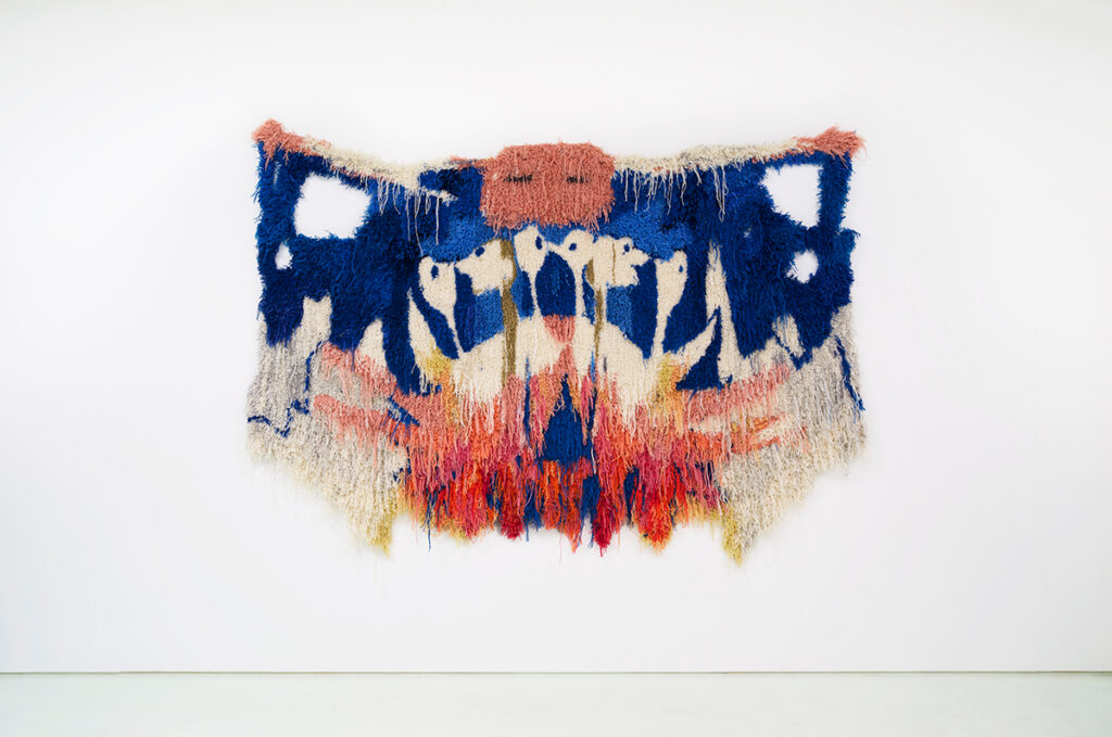 A multicolored tufted wall hanging resembling a rorschach test and featuring four mirrored holes within the organic shape of the tufting, exposing the wall behind the artwork.