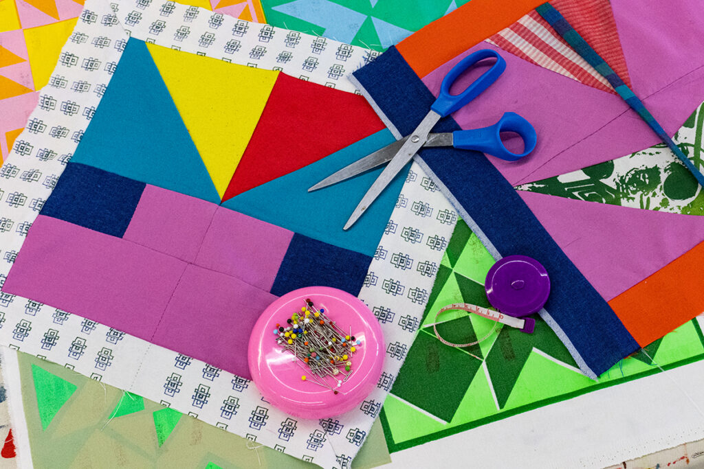 An image of a geometric patterned and multi-colored quilt square surrounded by measuring tape, scissors, and fabric pins.