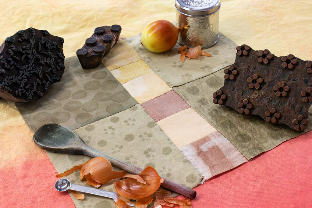 An image of a quilt square featuring a patchwork of different natural dye techniques in earthy colors. The square is surrounded by onion skins and wooden blocks for block printing.