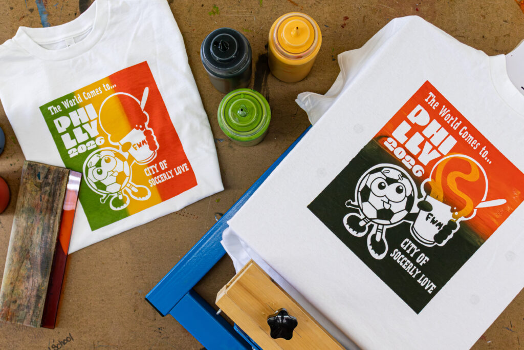 Two white t-shirts side by side on a table. One is folded and one is stretched over a t-shirt printing press. They both feature the same illustration of an anthropomorphized soccer ball holding a cup of water ice and celebrating the World Cup.