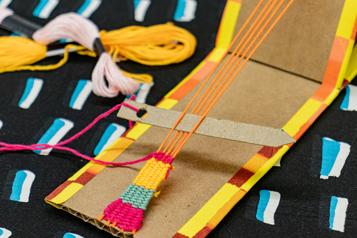 A small loom constructed of a folded cardboard sheet. Two ends of the cardboard are held in tension by stretched threads of yarn.