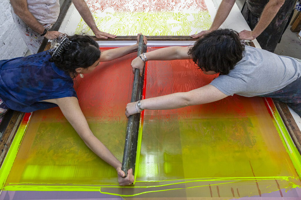A birds-eye view of two people standing on opposite sides of a screen, passing the squeegee to create a large-scale screenprint on fabric.