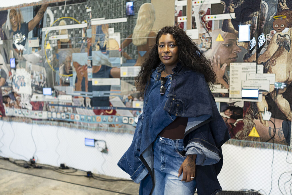The artist Qualeasha Wood, a Black woman wearing a fashionable denim outfit, poses for her portrait in front of a large tapestry.