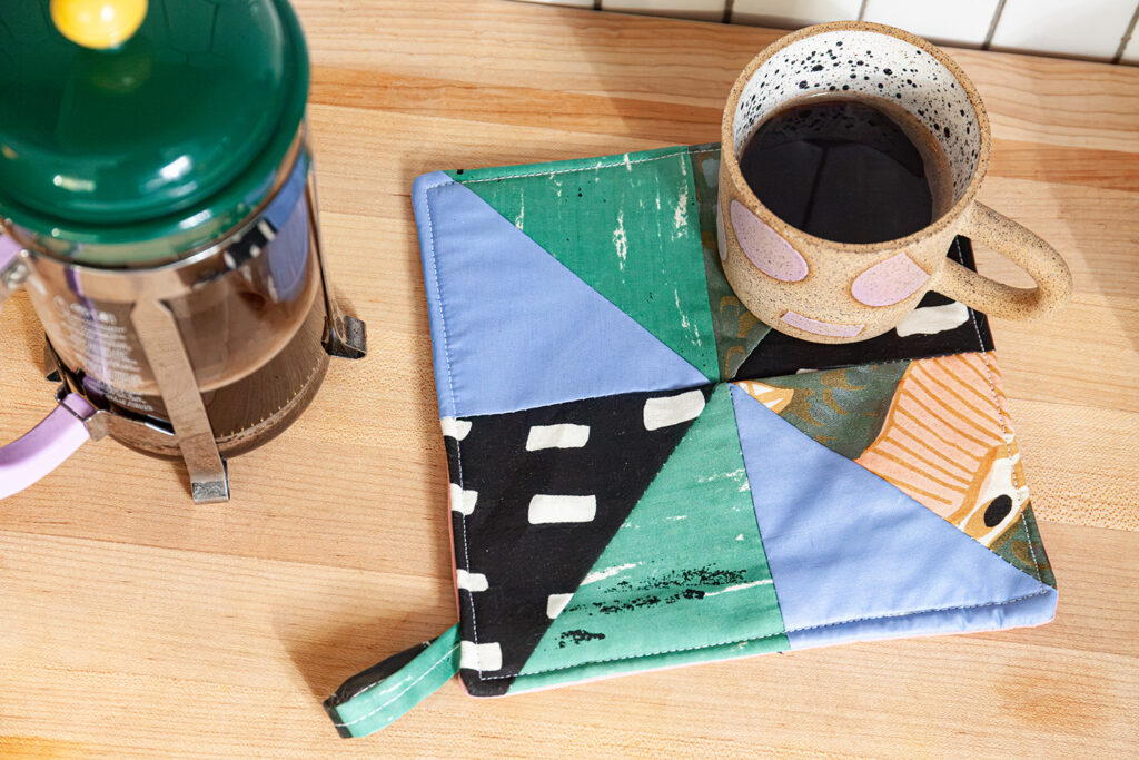 A square potholder sewn using different triangles of patterned fabrics. There is a ceramic coffee mug sitting on top of the potholder and a french press off to the left side.
