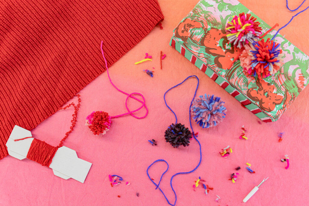 A photograph of a red sweater being unraveled and wound, then implied that the same yarn is used to create decorative pom poms. An example is shown of pom poms being used for gift wrap decoration.