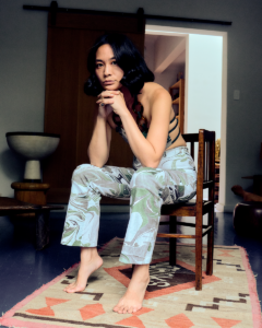 A portrait of the writer and curator Su Wu seated in a chair in a studio. She is leaning forward with her chin resting on her hands and her elbows planted on her thighs.